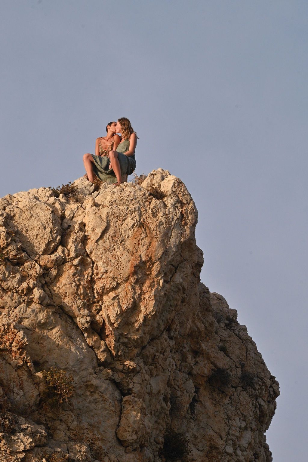 People sit atop a rugged limestone cliff formation against a blue sky at sunset.