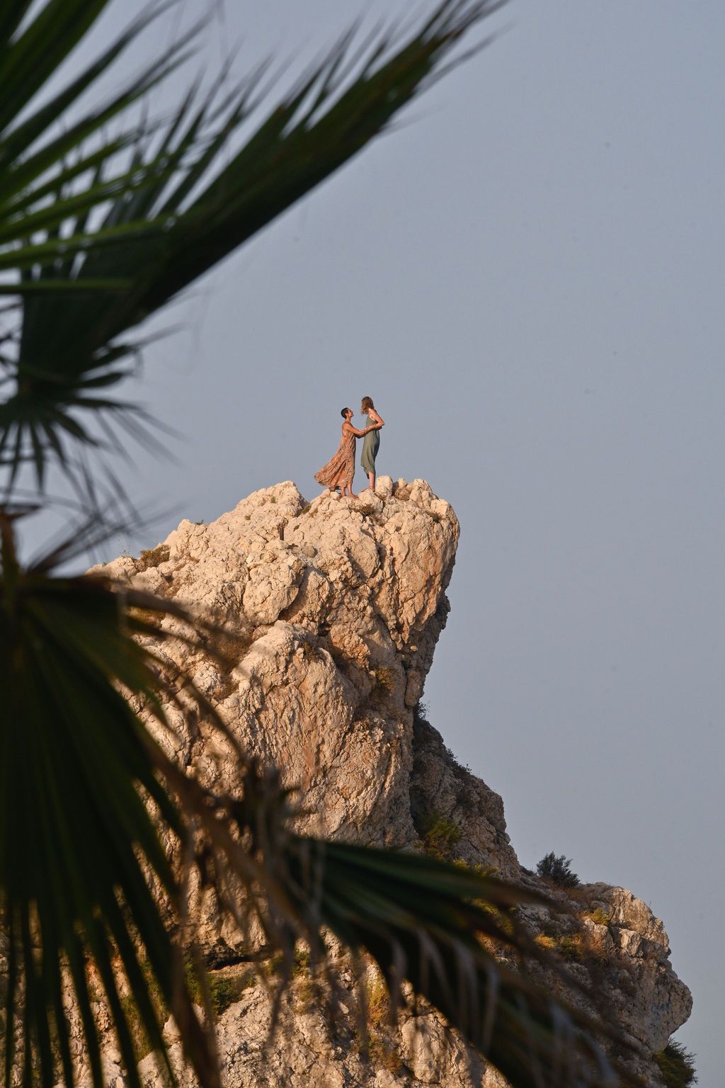 Jagged rock formation photographed at sunset with palm fronds silhouetted in the foreground against a grey sky.