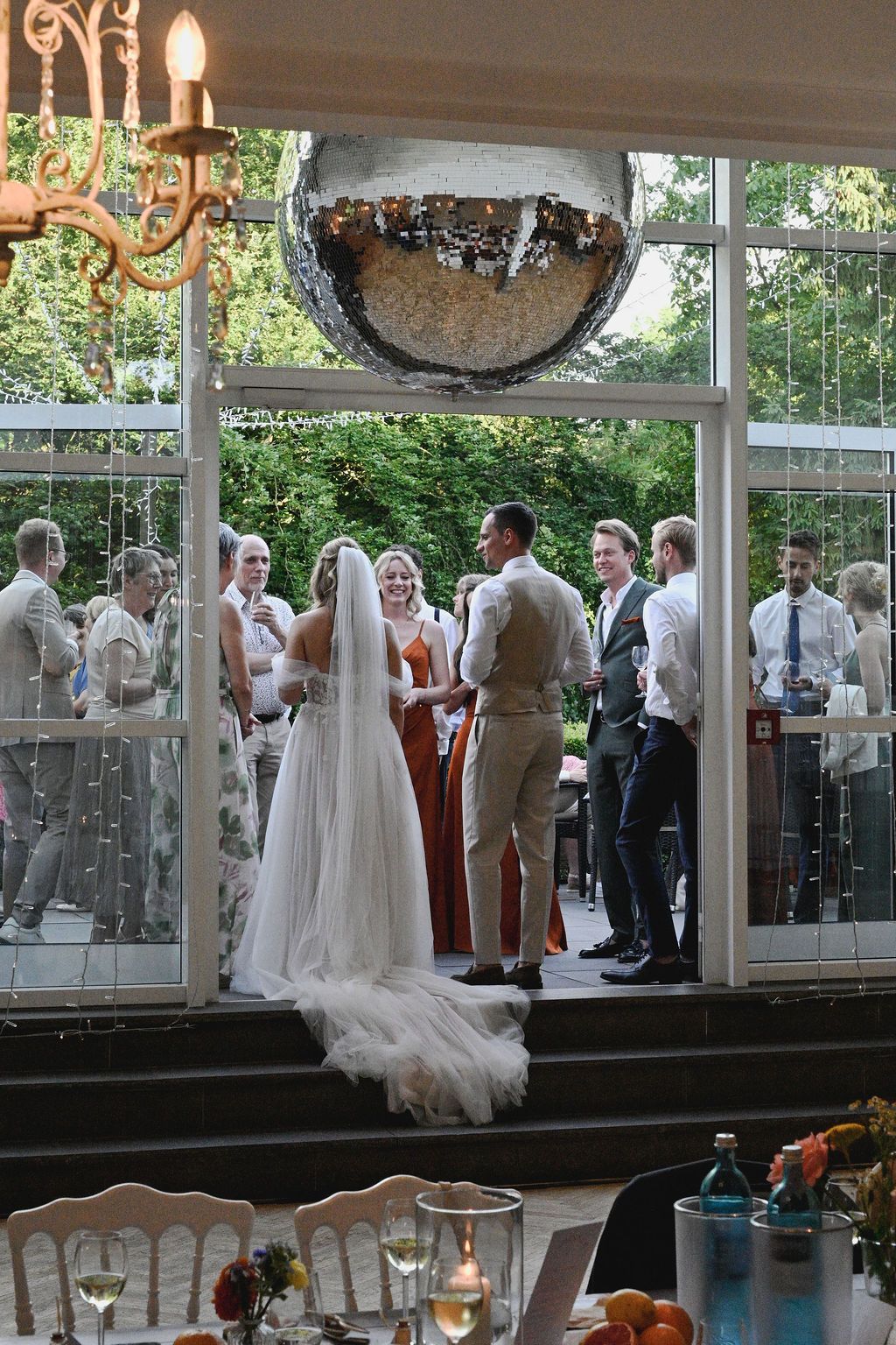 Wedding reception guests gather in an elegant glass-enclosed venue with white dresses and formal attire visible.