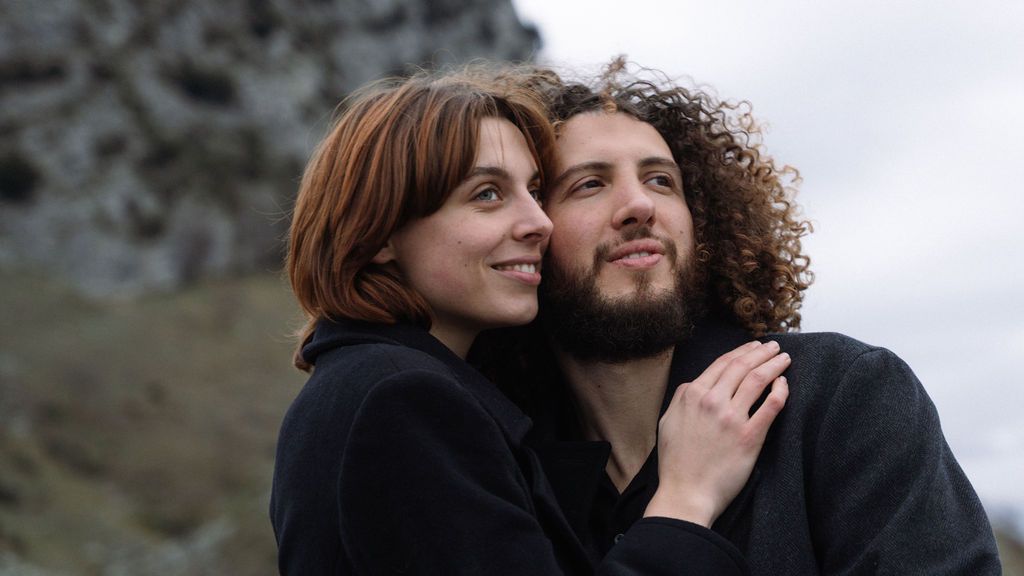 A young couple embracing outdoors with rocky terrain behind them, woman with red hair smiling warmly.