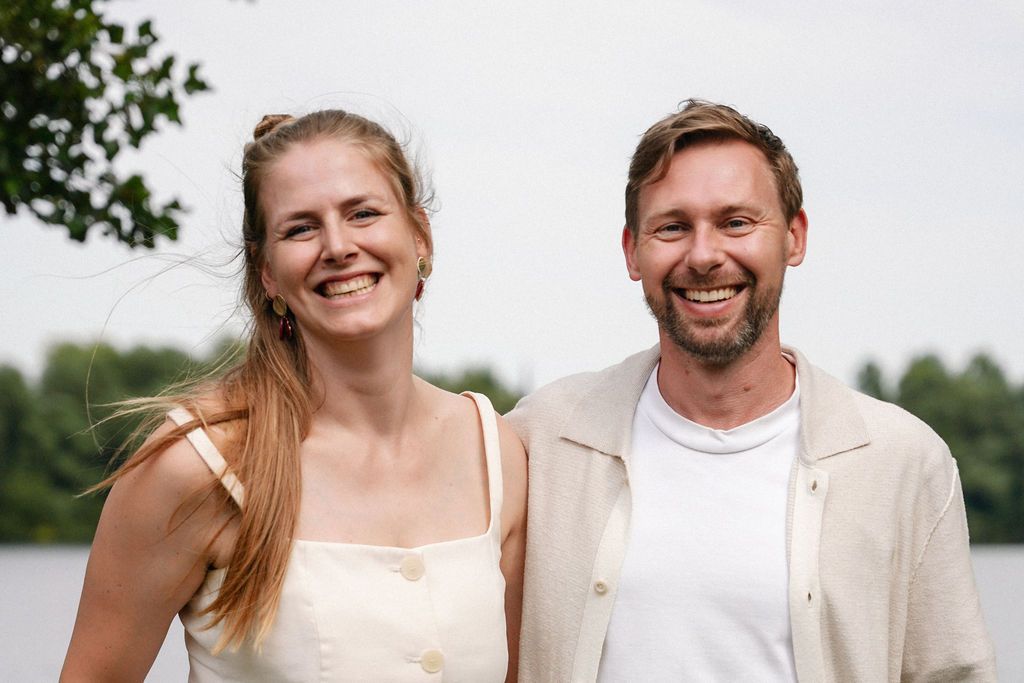 A happy couple wearing light colored clothing smiles together outdoors by a lake on a summer day.