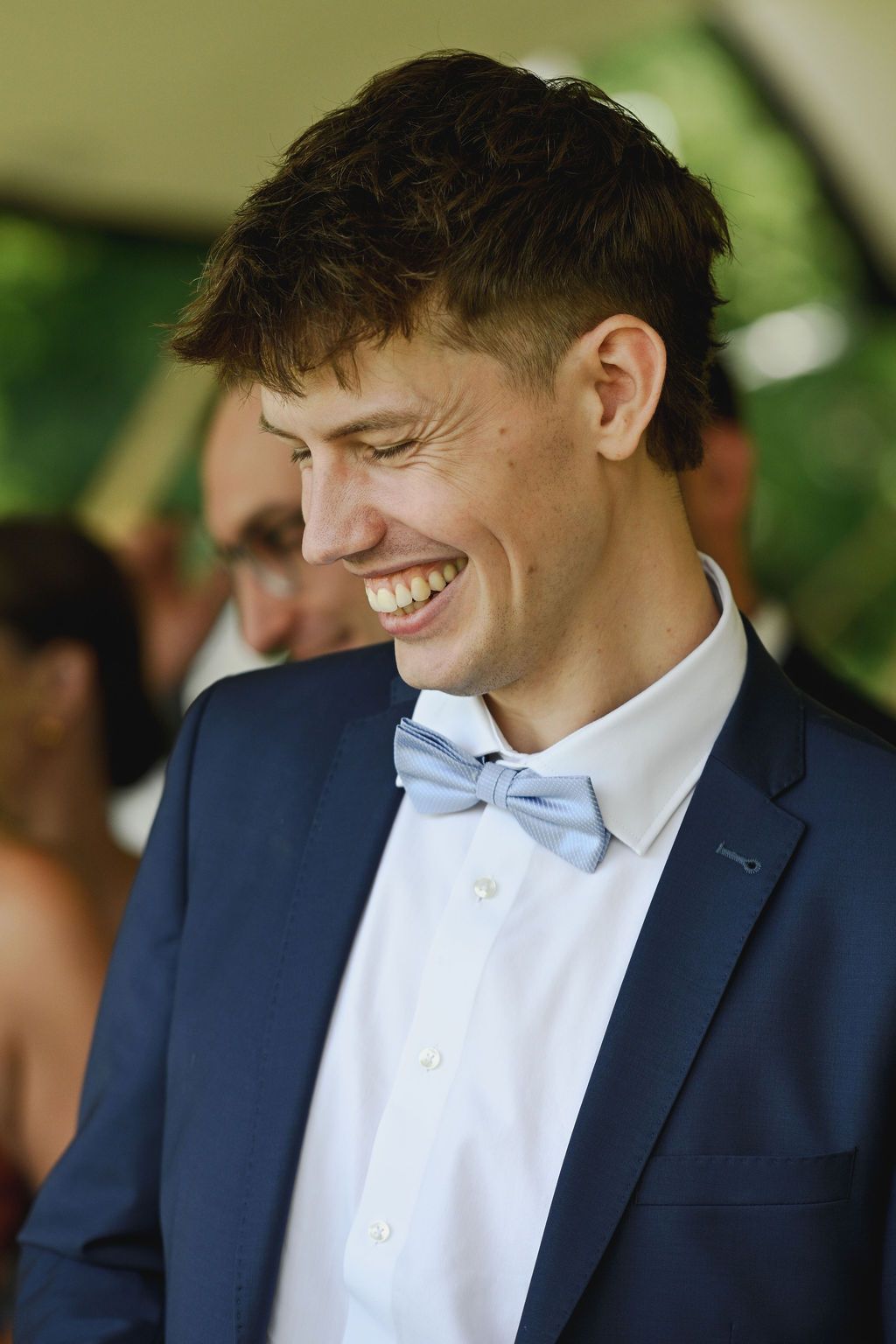 A person in a navy suit and light blue bowtie smiles warmly during a formal occasion.