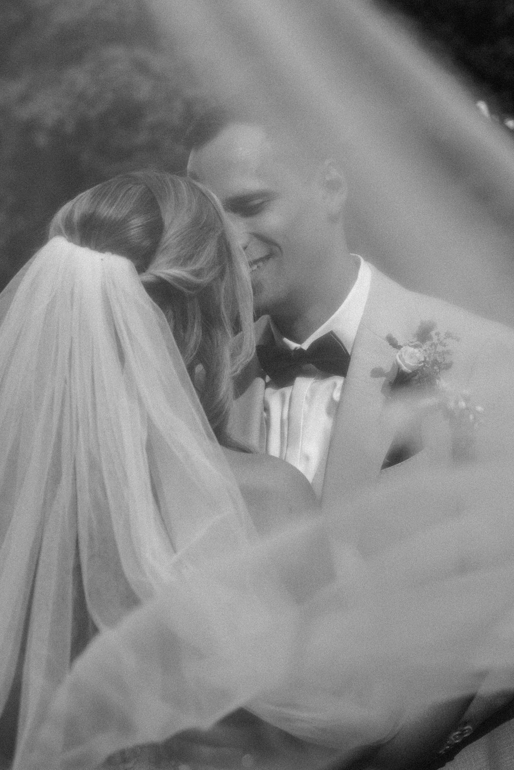 A romantic black and white wedding photo showing a tender moment between newlyweds through a flowing veil.