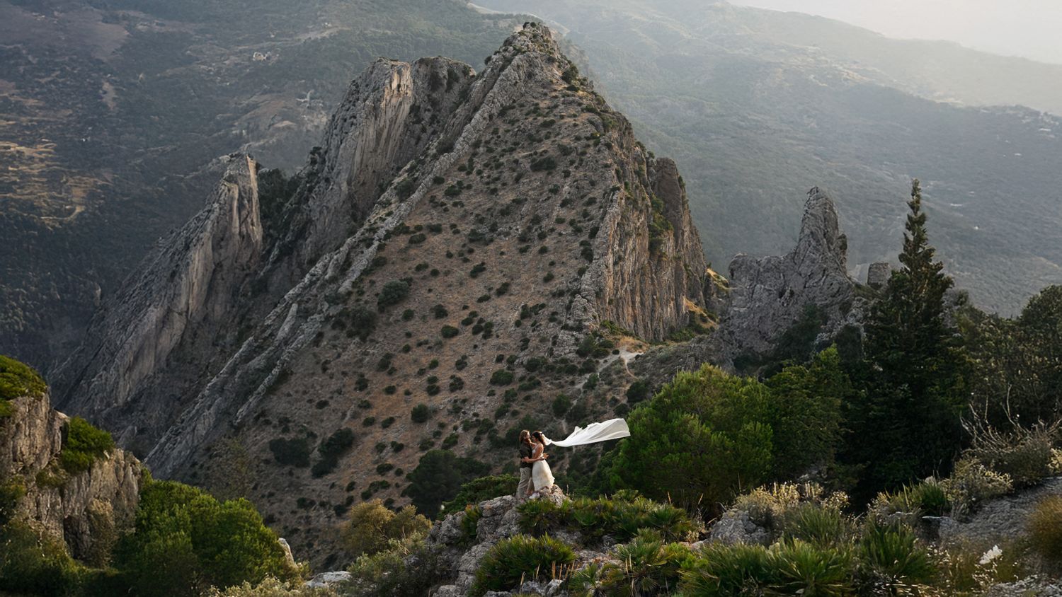 Bride in white dress with flowing veil stands on rocky mountain ridge with dramatic cliffs and valley views.