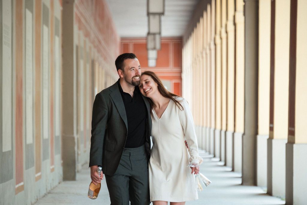 Happy couple walking arm in arm through elegant columned arcade, woman in white dress, man in dark suit.