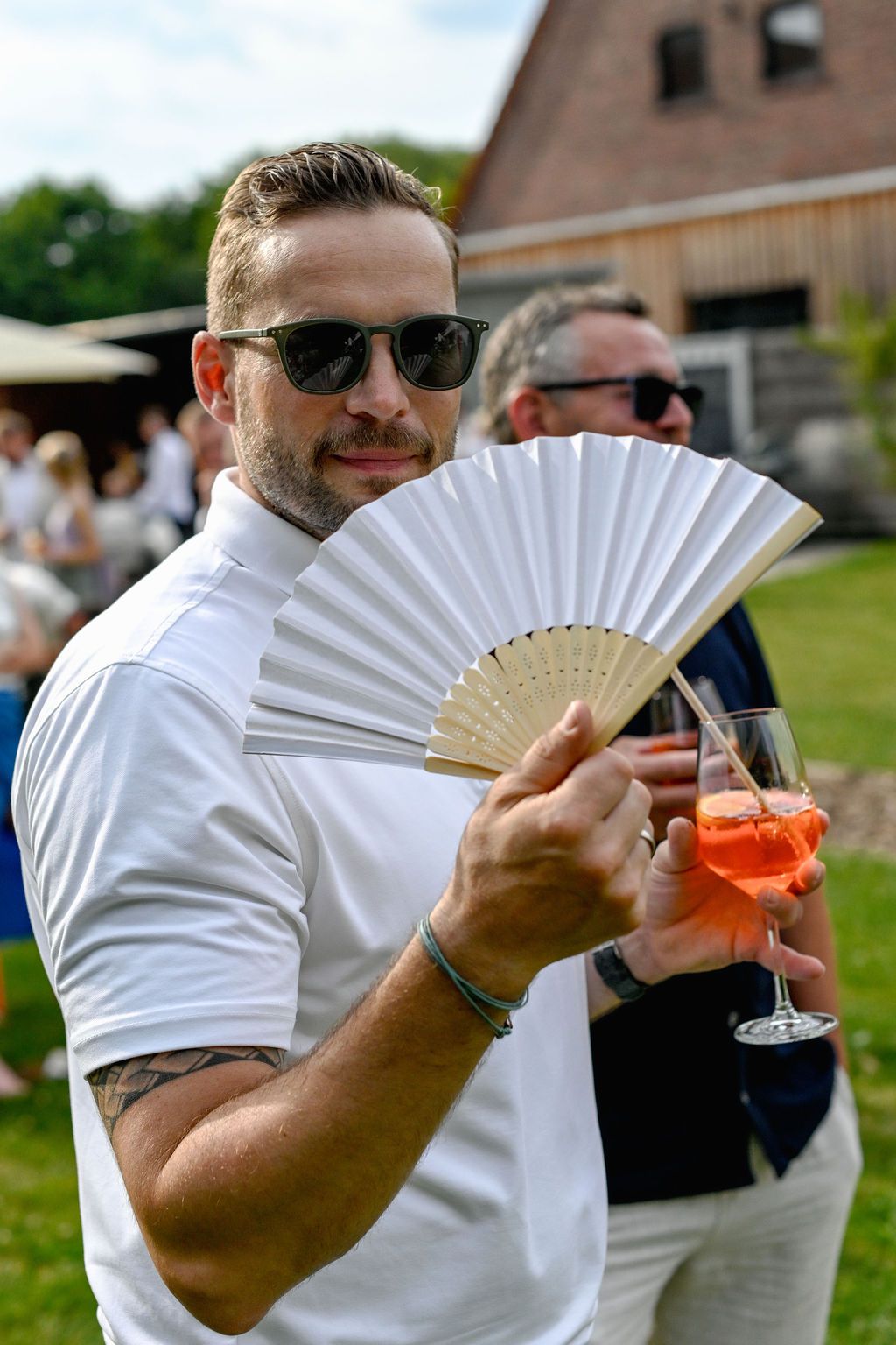 A person in a white shirt holding a decorative fan and cocktail glass poses playfully at an outdoor event.