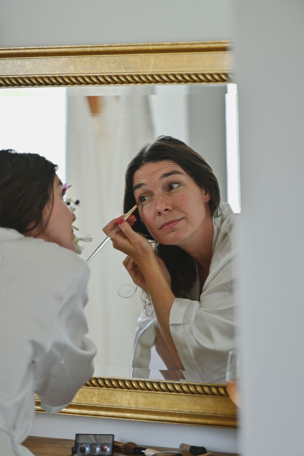 Person in white robe applies makeup while looking in an ornate gold-framed mirror.