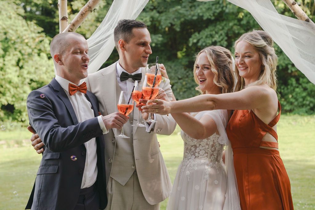 Wedding party members raise their glasses in a celebratory toast outdoors during a summer celebration.