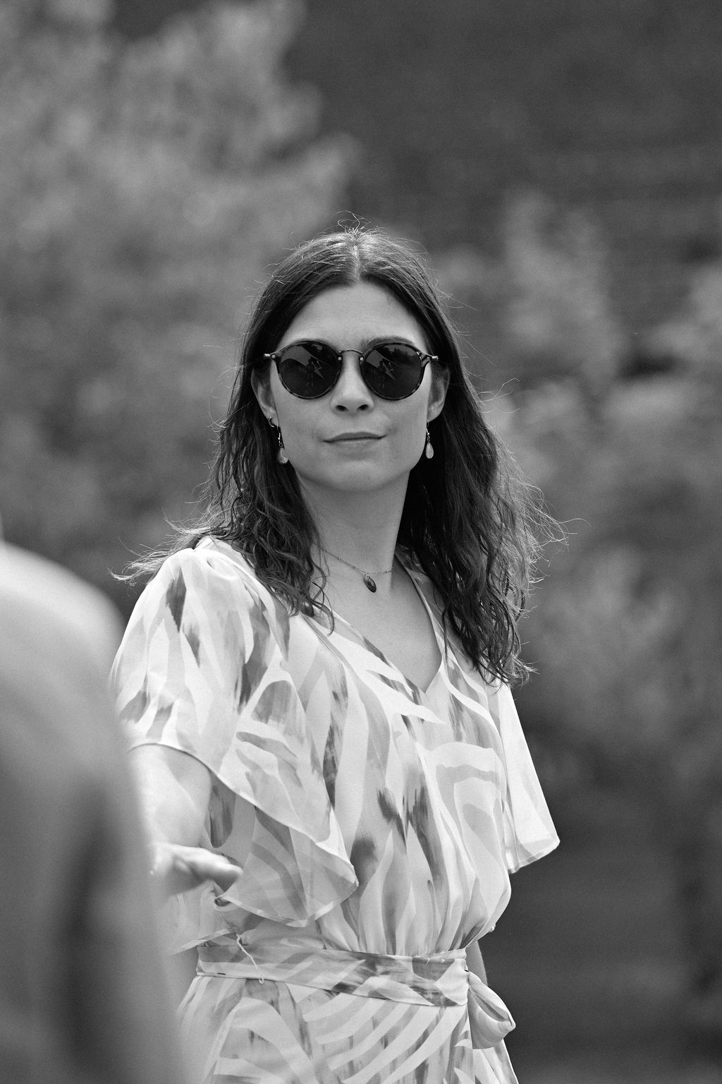 A person in a white ruffled dress and sunglasses poses outdoors in a black and white photograph.
