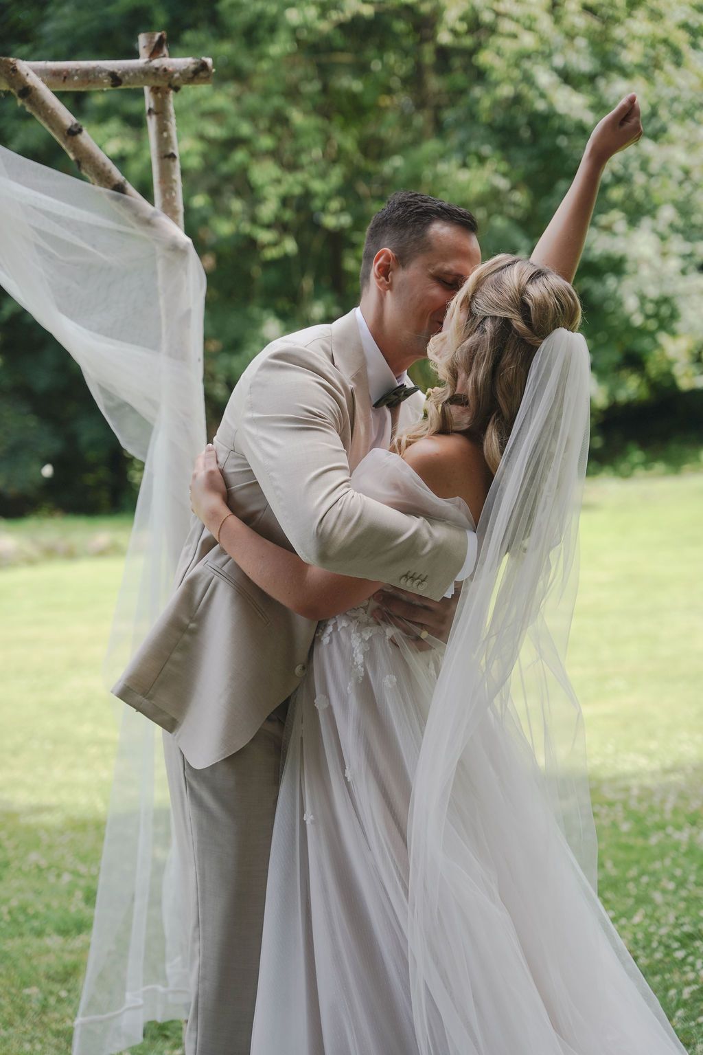 A romantic wedding couple shares a tender kiss in a garden setting while the bride's veil flows in the breeze.