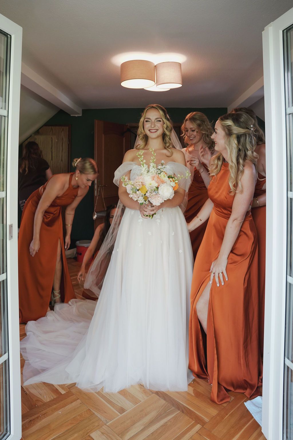 A wedding party in orange bridesmaid dresses gathers around a bride in a white gown in a bright sunlit room.