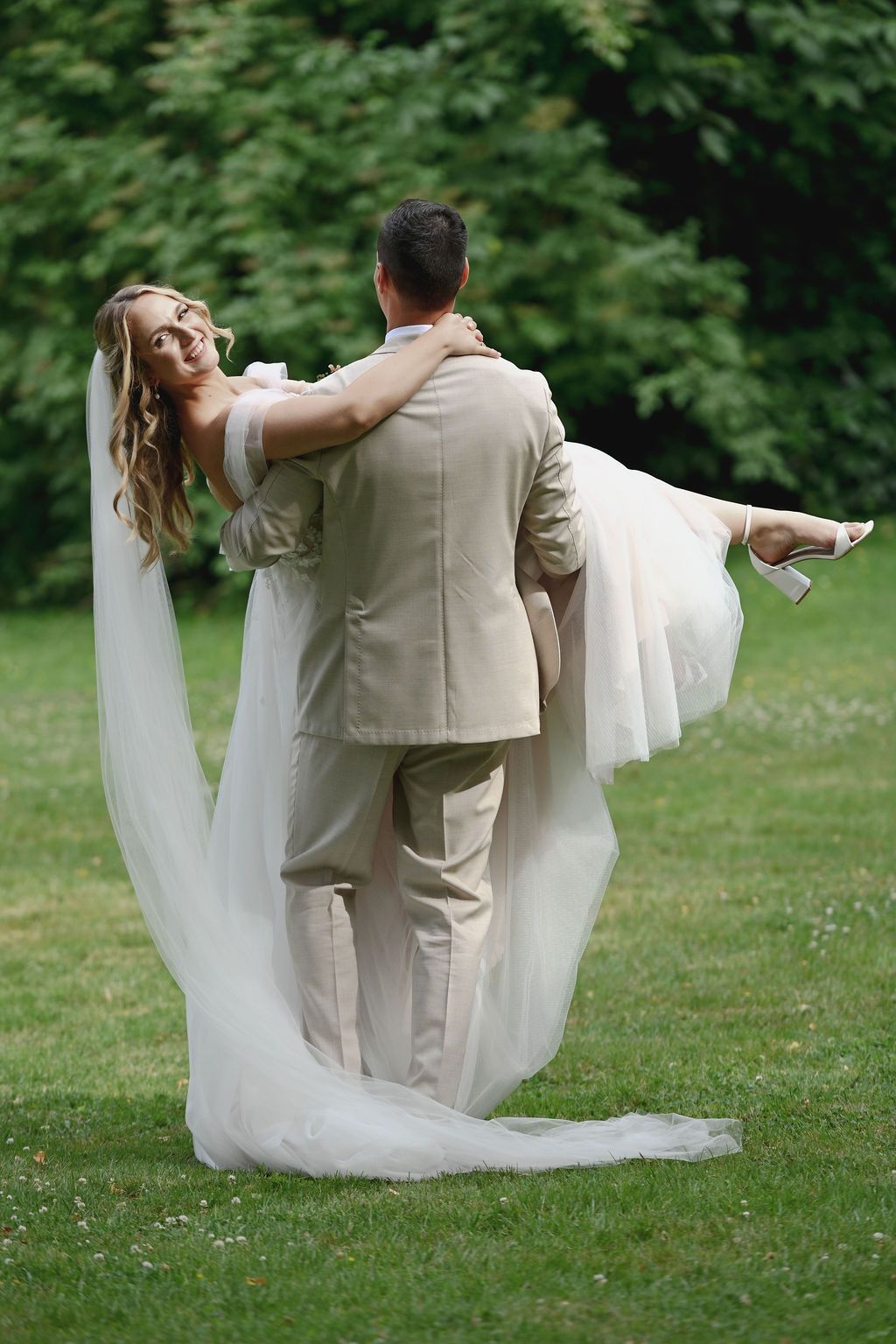 A couple in wedding attire dances playfully on a green lawn surrounded by trees on their special day.