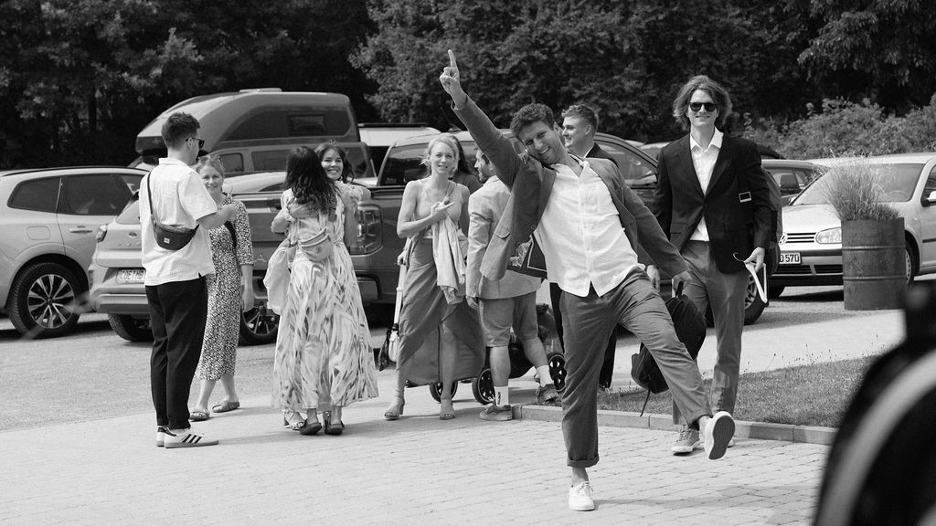Black and white panoramic image showing a crowd of people walking outdoors near parked cars on a sunny day.