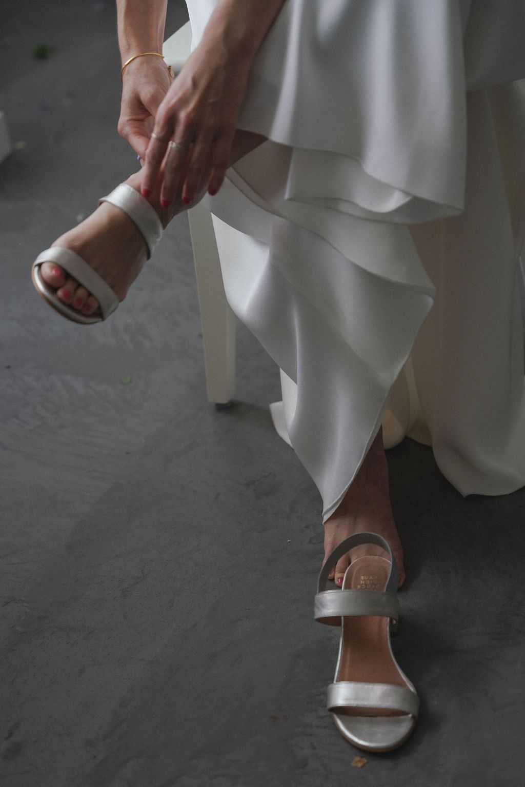 Bride adjusting silver strappy heeled sandals beneath a layered white wedding gown while seated.