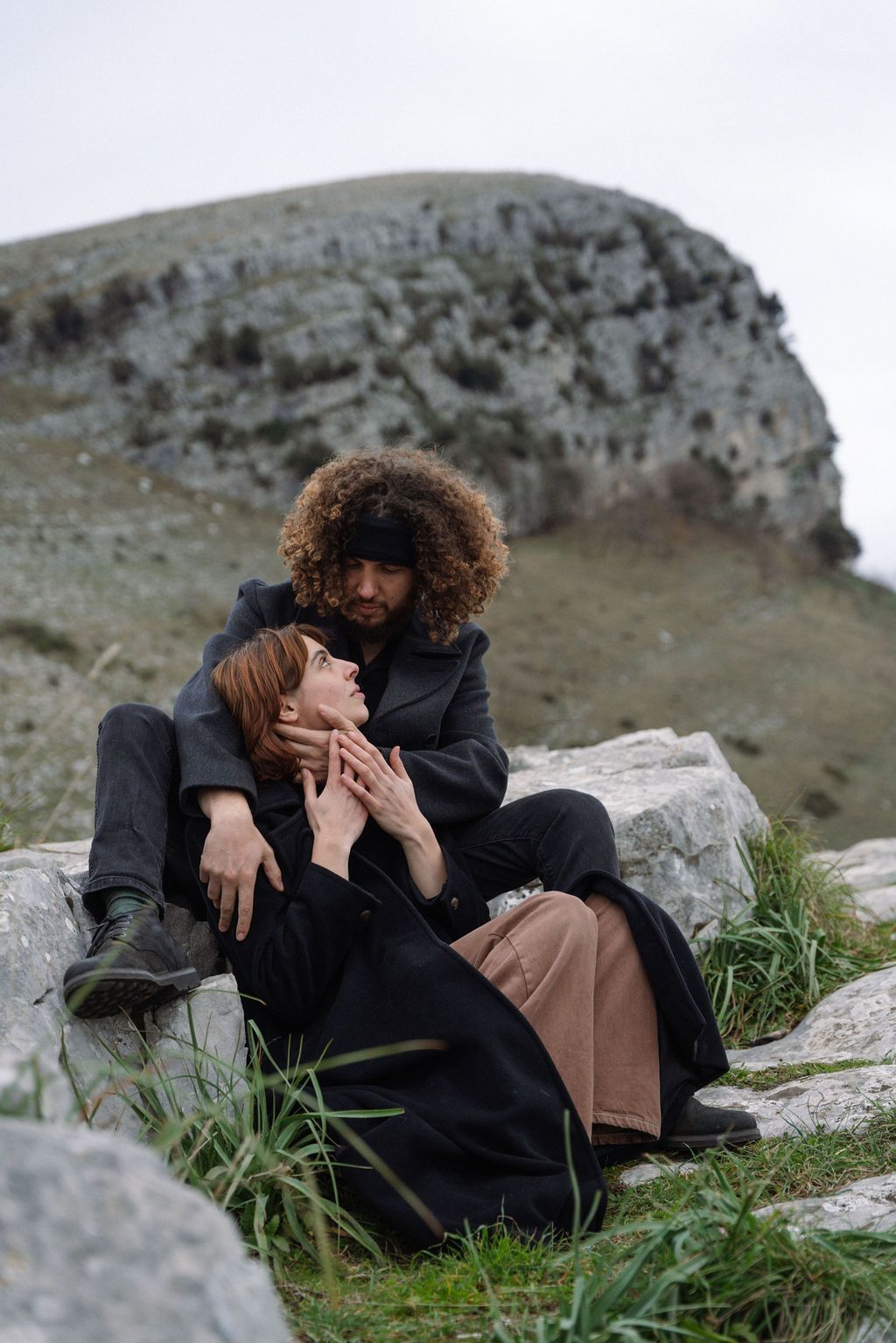 Two people in dark winter clothing embrace while sitting on rocky terrain with a mountainous landscape in the background.