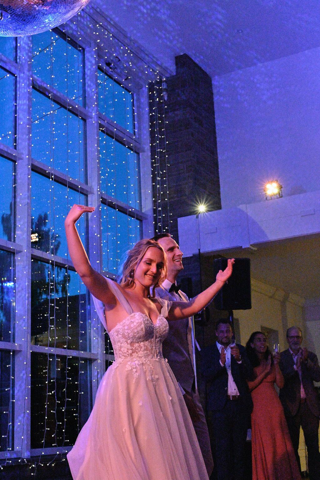 Wedding couple and guests dance together under blue lighting and string lights at an evening reception celebration.