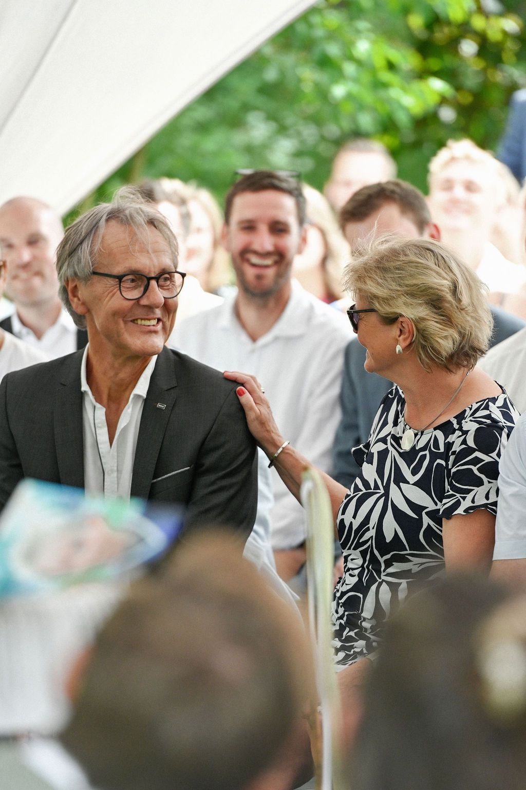 A social gathering shows people smiling and interacting at an outdoor event, with guests wearing casual formal attire.