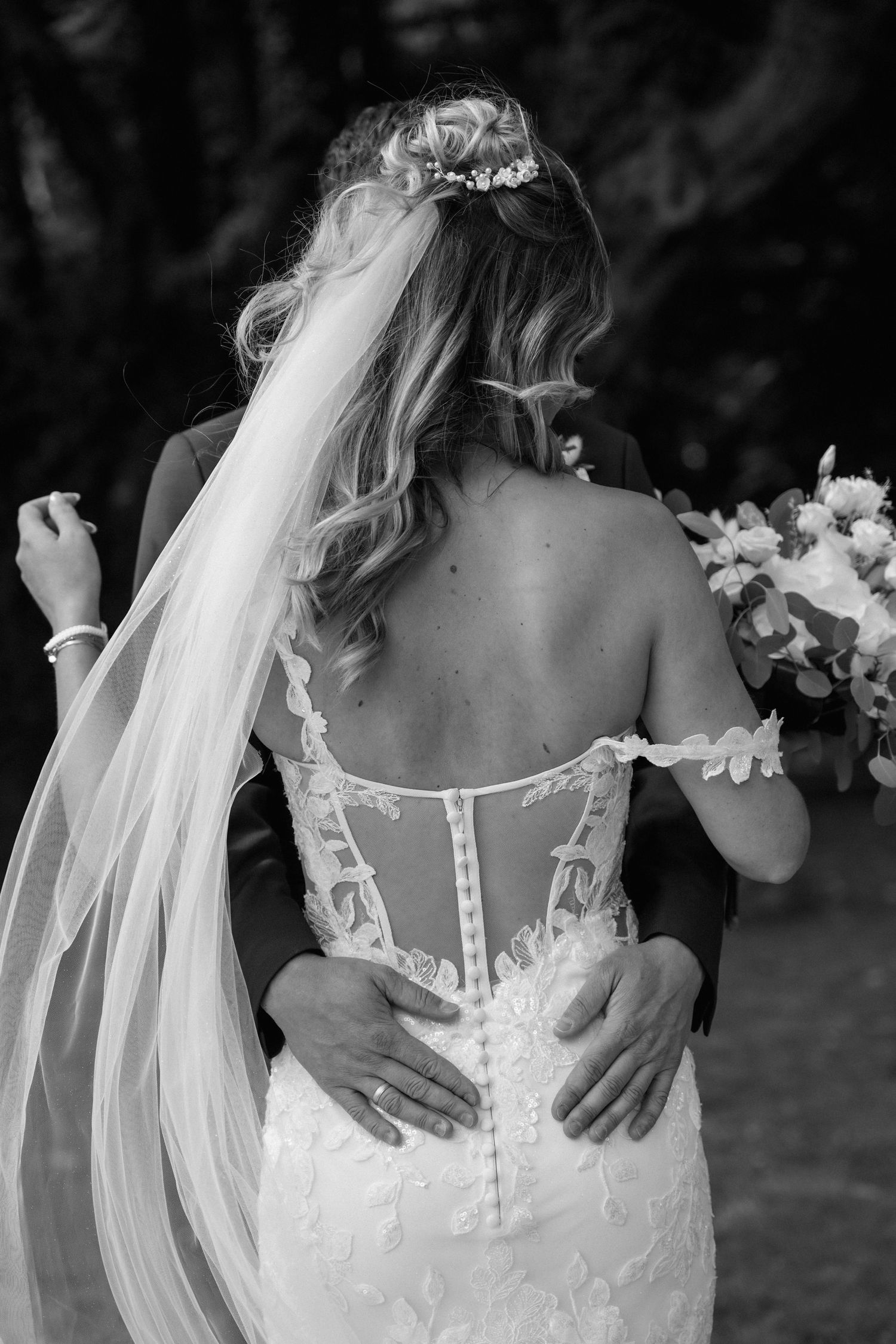 Black and white wedding photo showing bride in strapless lace gown with long veil and hands embracing from behind.