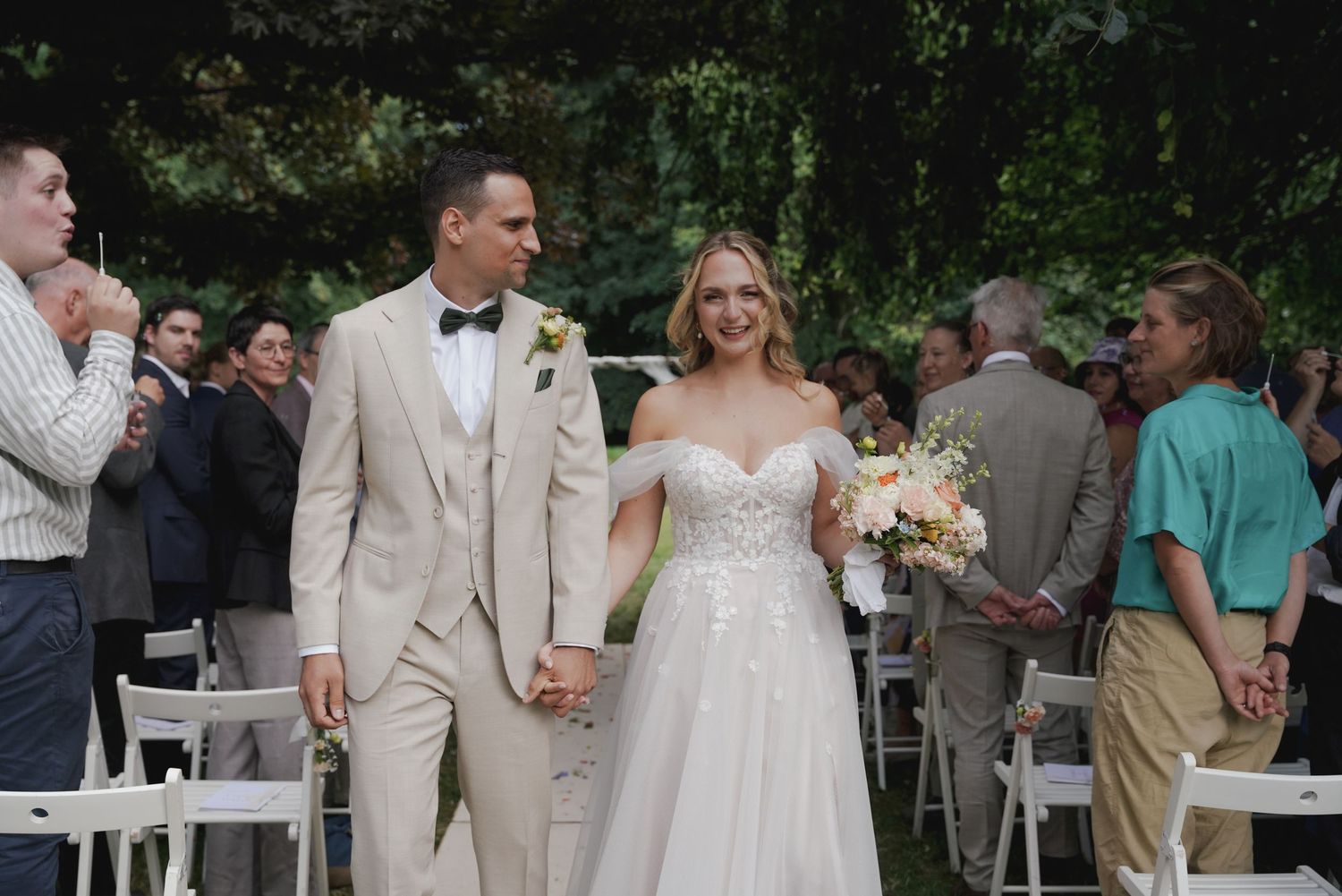 A couple walks hand in hand at an outdoor wedding ceremony with white chairs and guests in the background.
