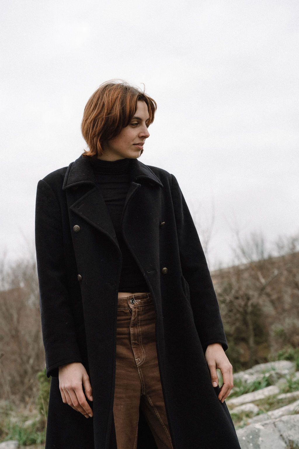 A person wearing a black wool coat and brown pants poses against a natural outdoor backdrop.