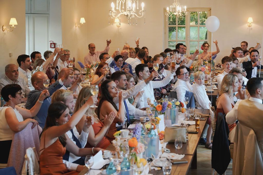 Wedding guests raise their glasses in a toast during an elegant reception with crystal chandeliers and soft lighting.