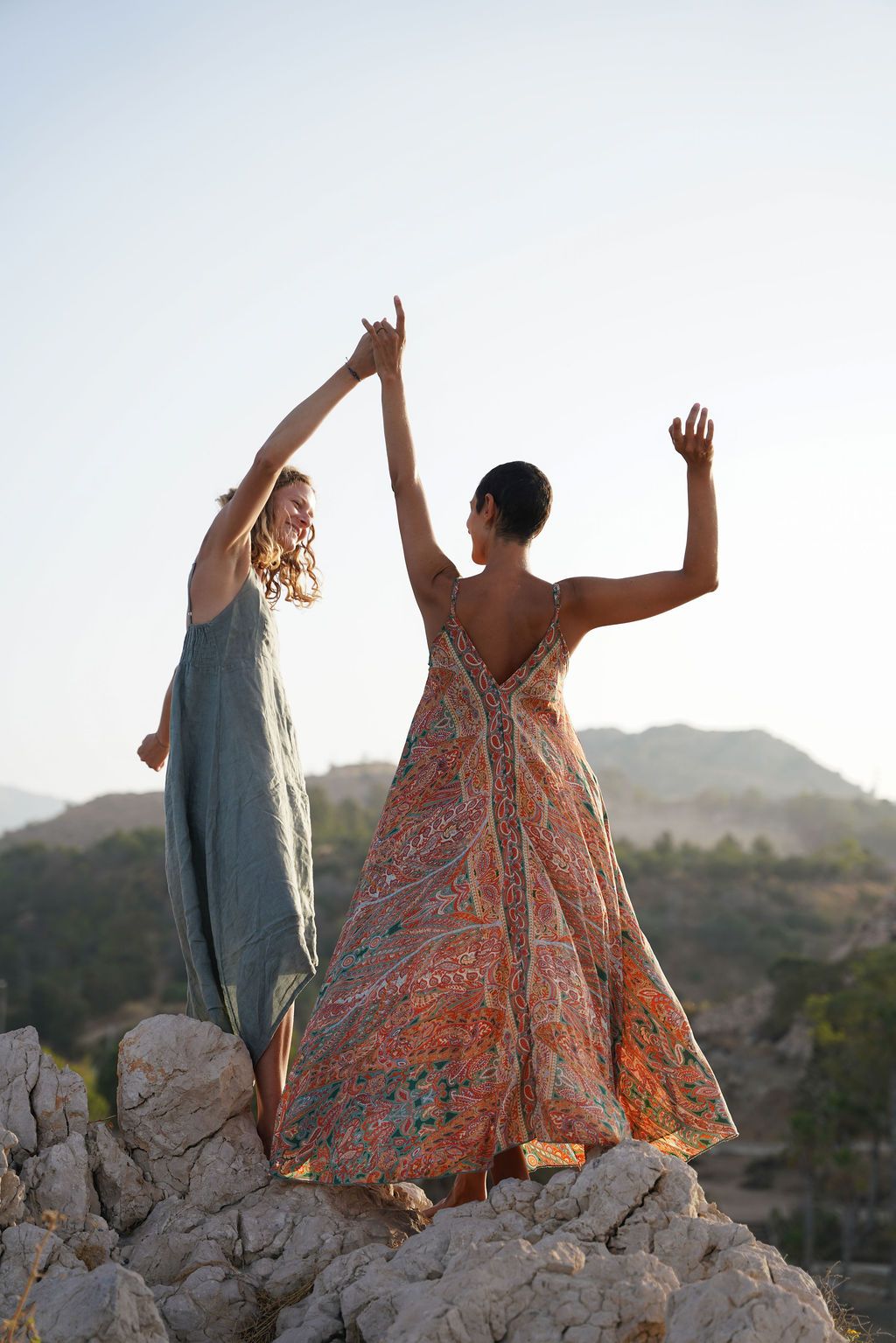 Two people stand on rocky terrain with raised arms at sunset, wearing flowing dresses in contrasting colors.