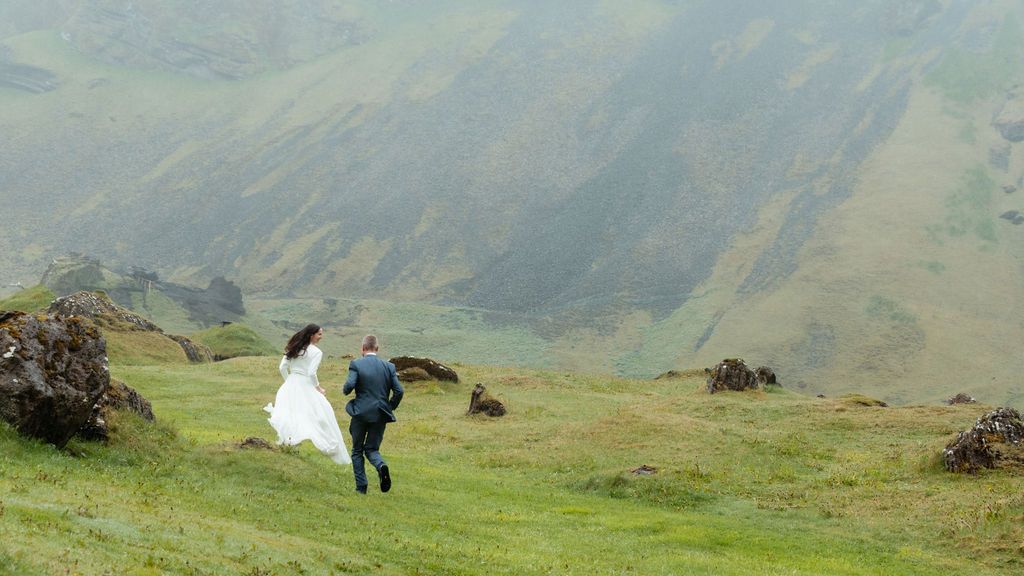 A bride and groom run hand in hand across a lush green hillside with misty mountains in the background.