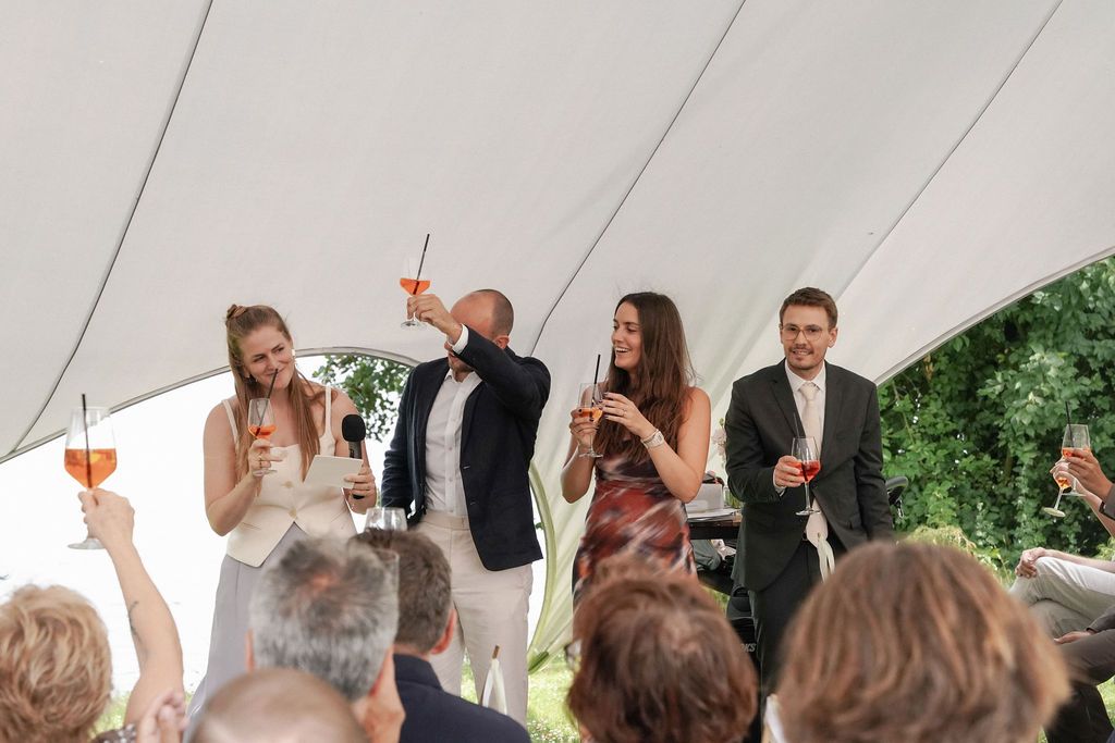 A group of wedding guests raise their glasses in a toast under a white tent during an outdoor celebration.