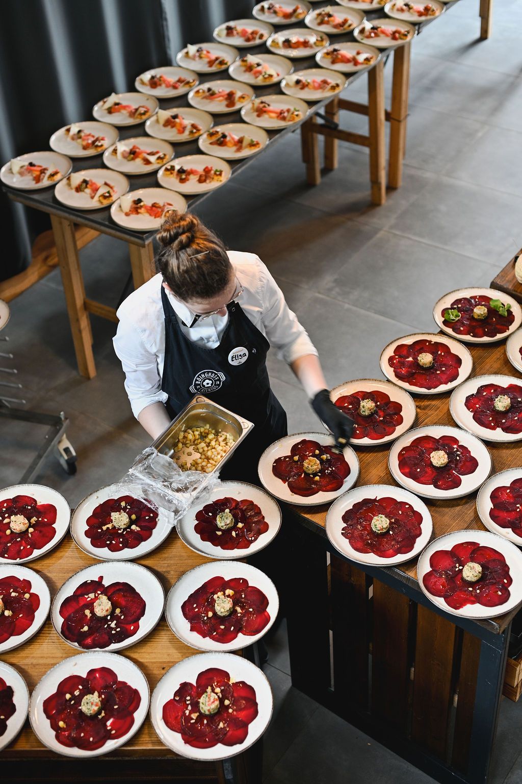 Overhead view of restaurant staff plating multiple dishes with vibrant red beet carpaccio on white scalloped plates.