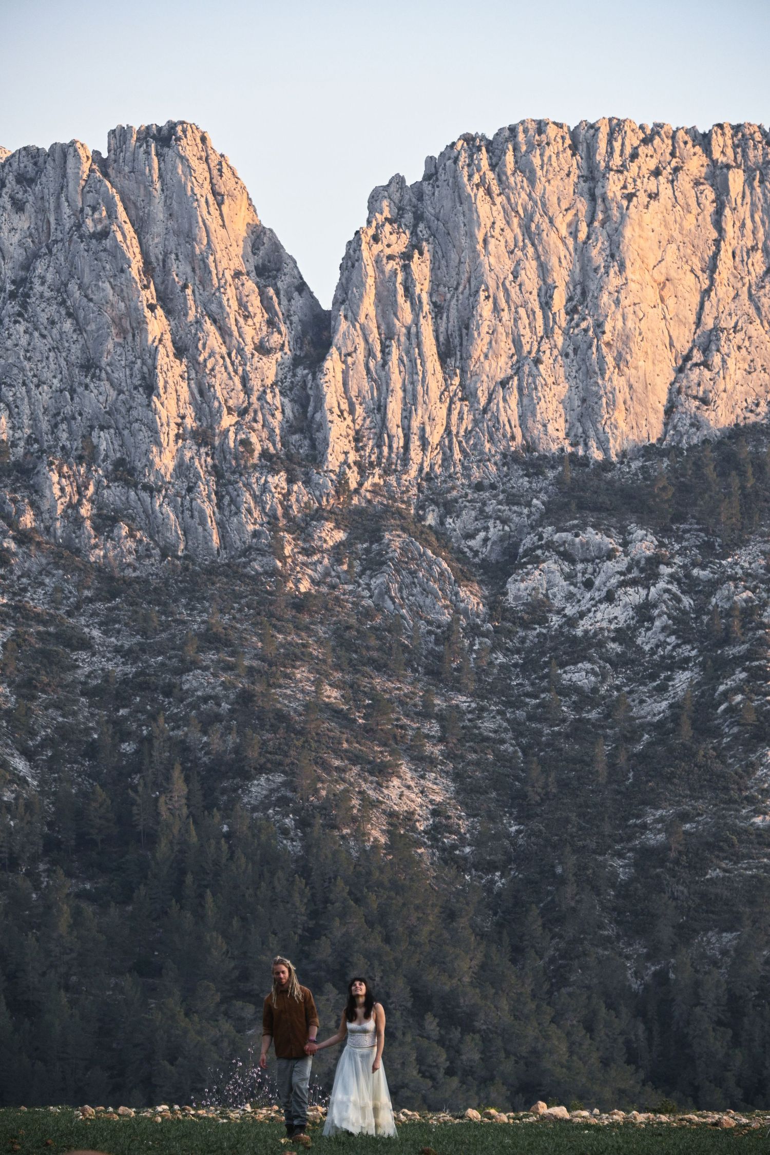 Dramatic mountain cliffs tower over couple standing in romantic pose against rugged landscape.