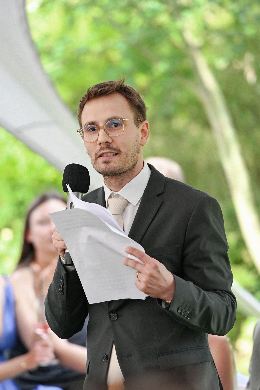 A speaker in a black suit gestures expressively while reading from papers at an outdoor event.