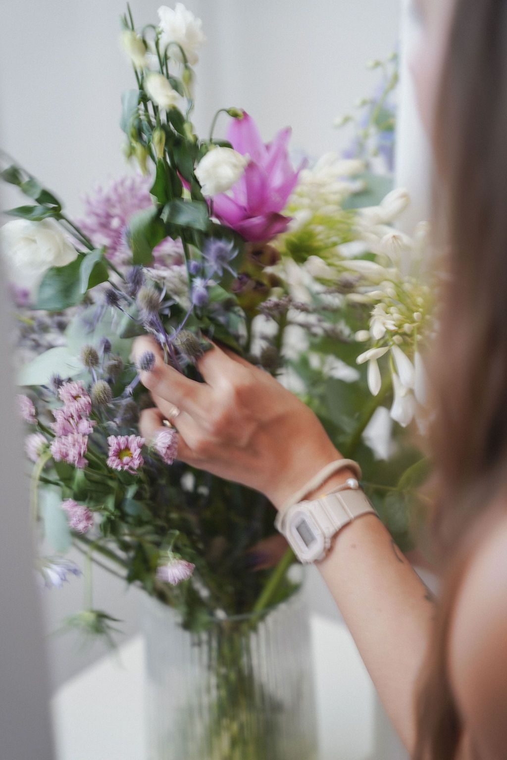 Hands arranging fresh pink and purple flowers in a glass vase against a white background.