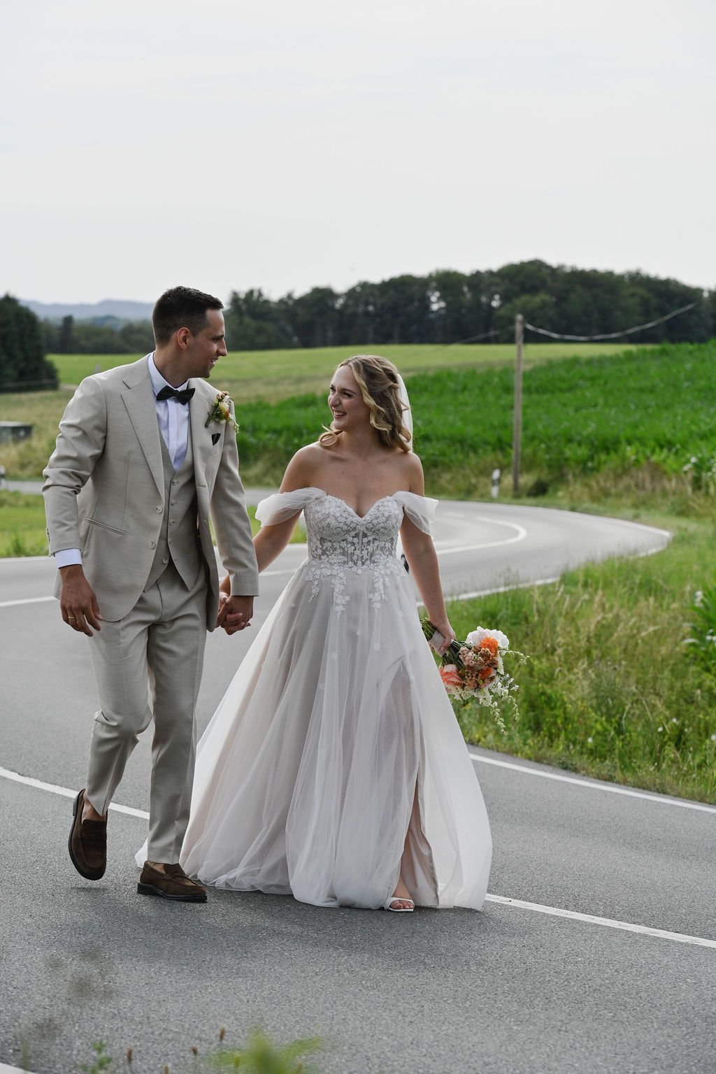 Newlywed couple walking and dancing together on a rural country road surrounded by green fields.