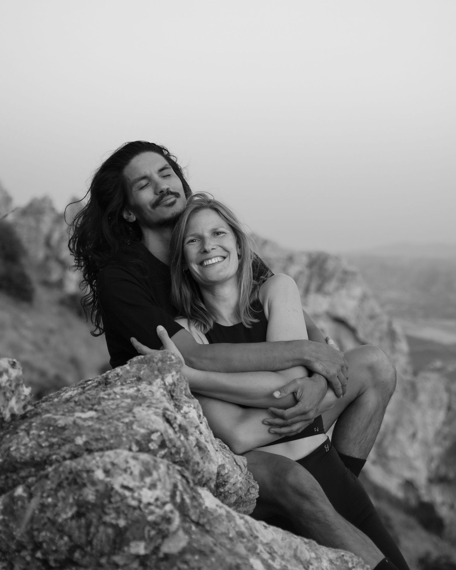 Two people share joyful embraces and laughter while sitting on rocky terrain in black and white photographs.