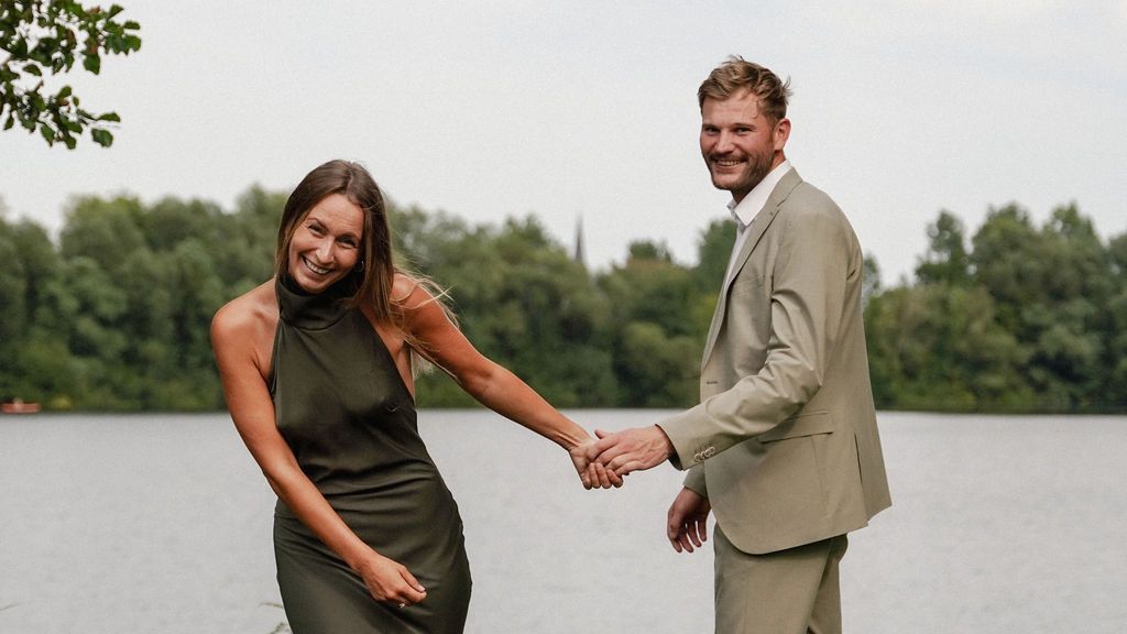 A smiling couple holds hands outdoors near a lake, woman in green dress, man in beige suit, lush trees behind them.