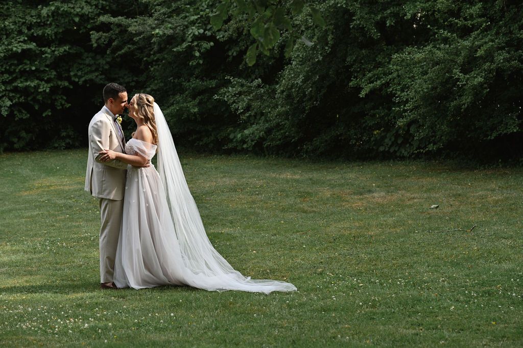 A couple in wedding attire embrace on a lush green lawn with forest backdrop during an outdoor ceremony.