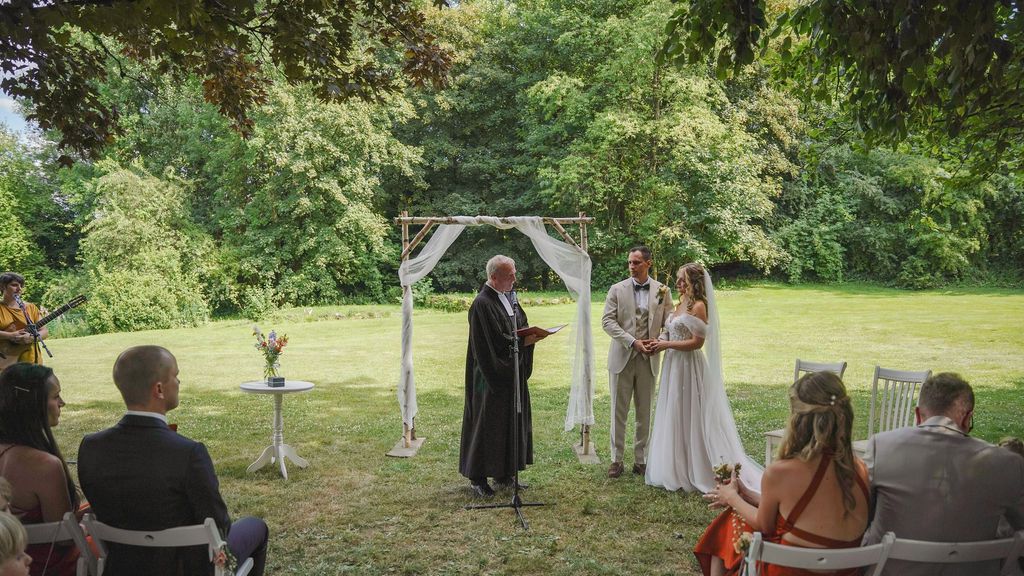 An outdoor wedding ceremony takes place in a lush green garden setting with guests seated on white chairs.