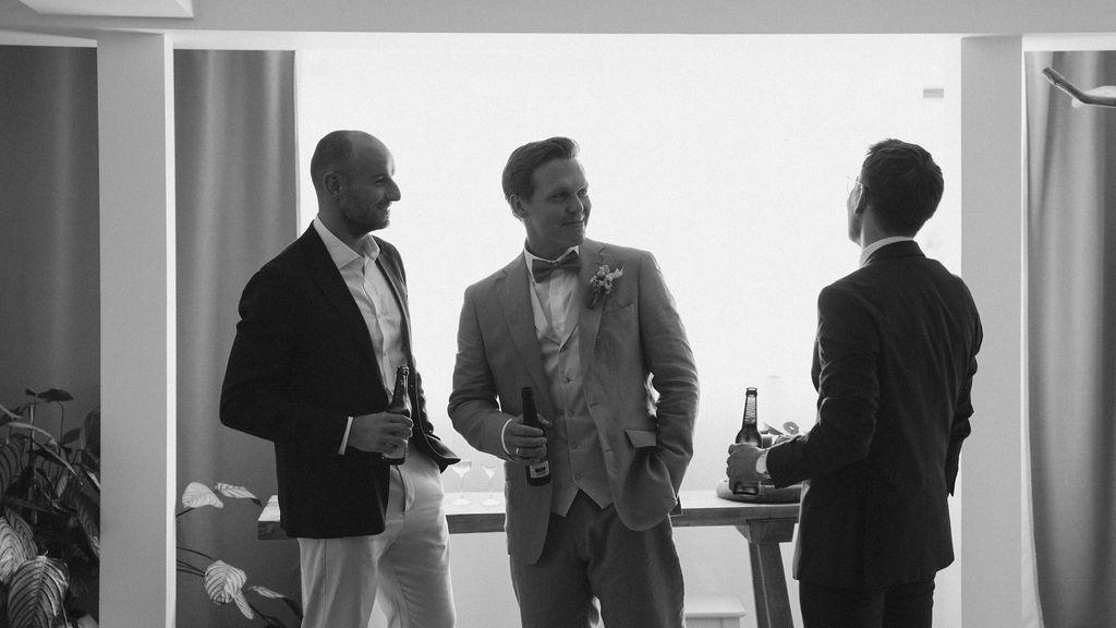 Black and white photo of three men in suits conversing at an elegant wedding reception with scenic views.