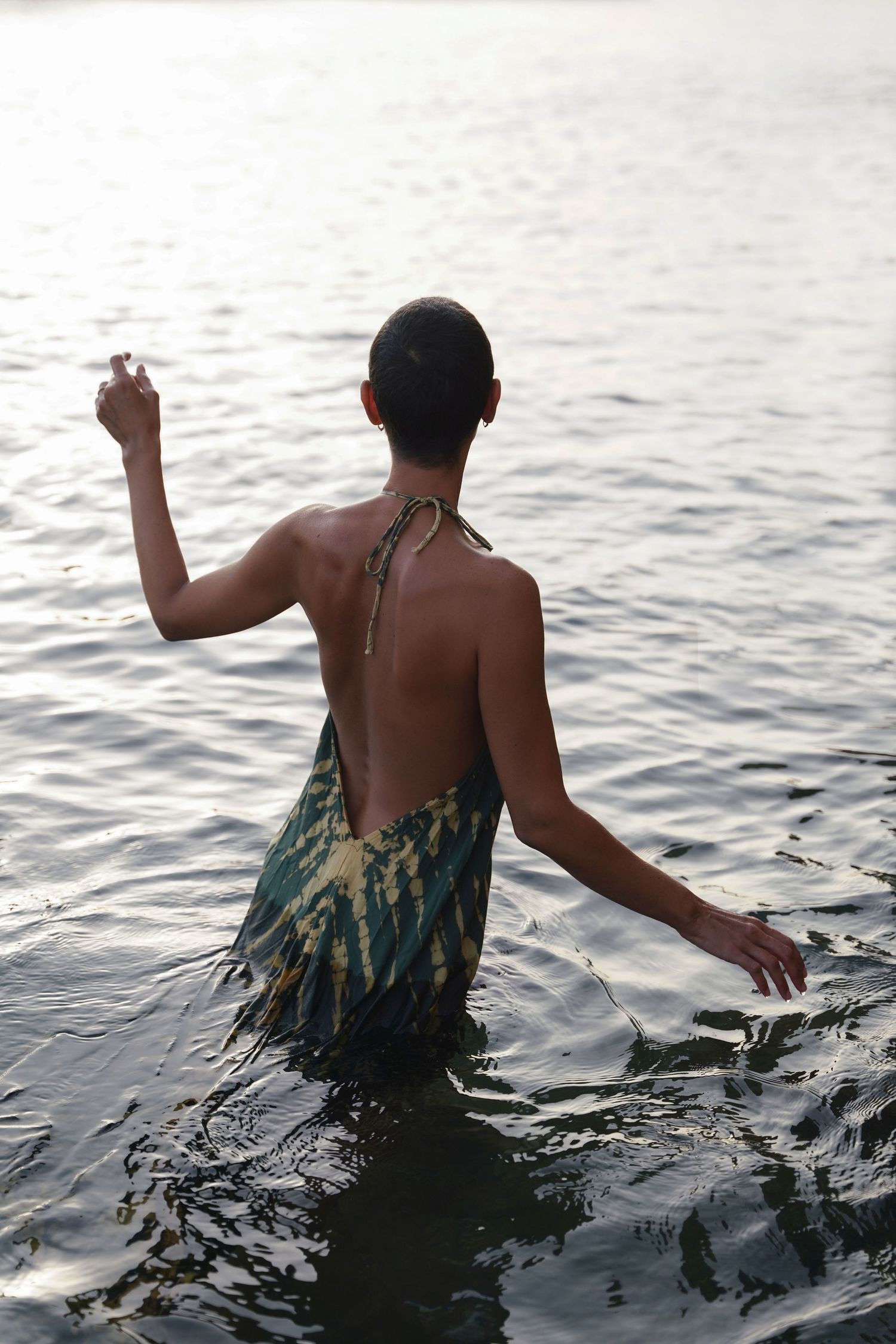 A silhouetted figure in tie-dye clothing wading in calm ocean waters at sunset.