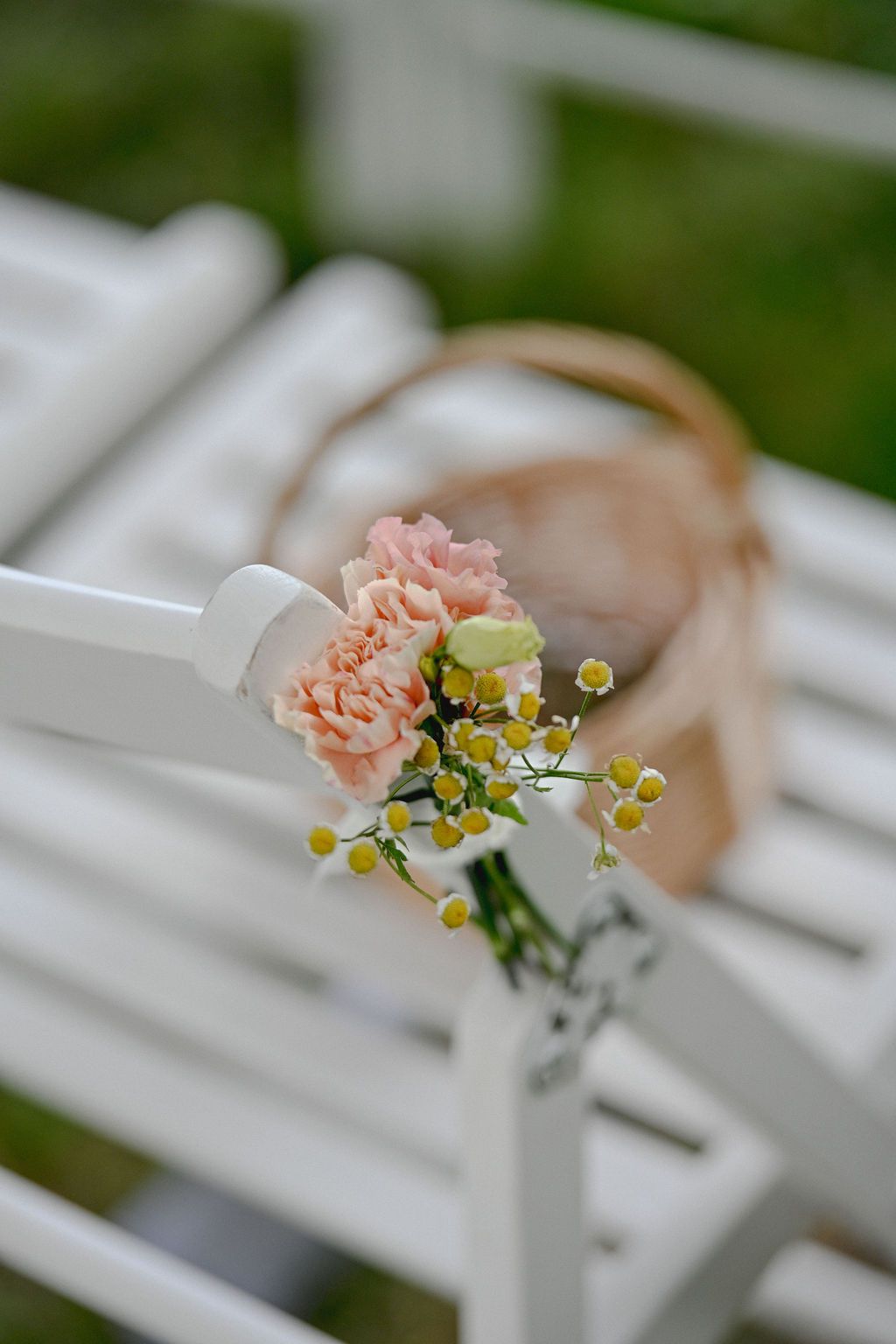 Delicate pink and yellow flowers decorate white chairs at an outdoor wedding ceremony.