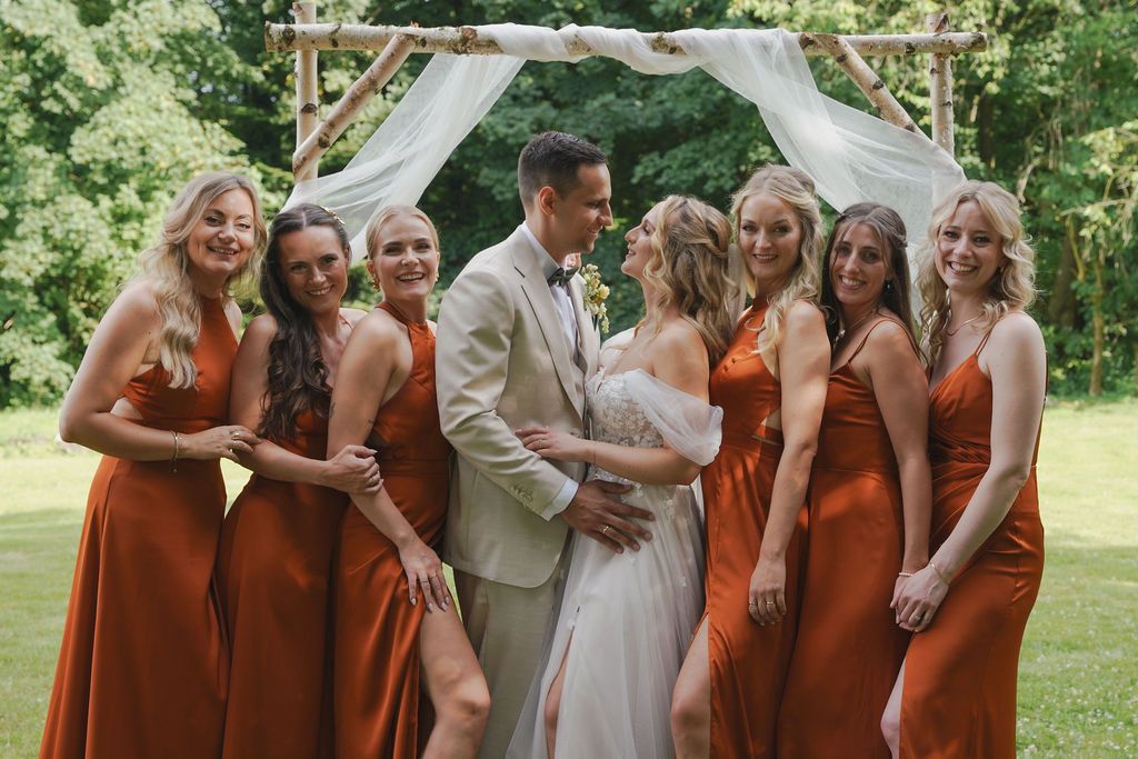 Wedding party poses outdoors with bridesmaids in rust-colored dresses under a white arch on a sunny day.