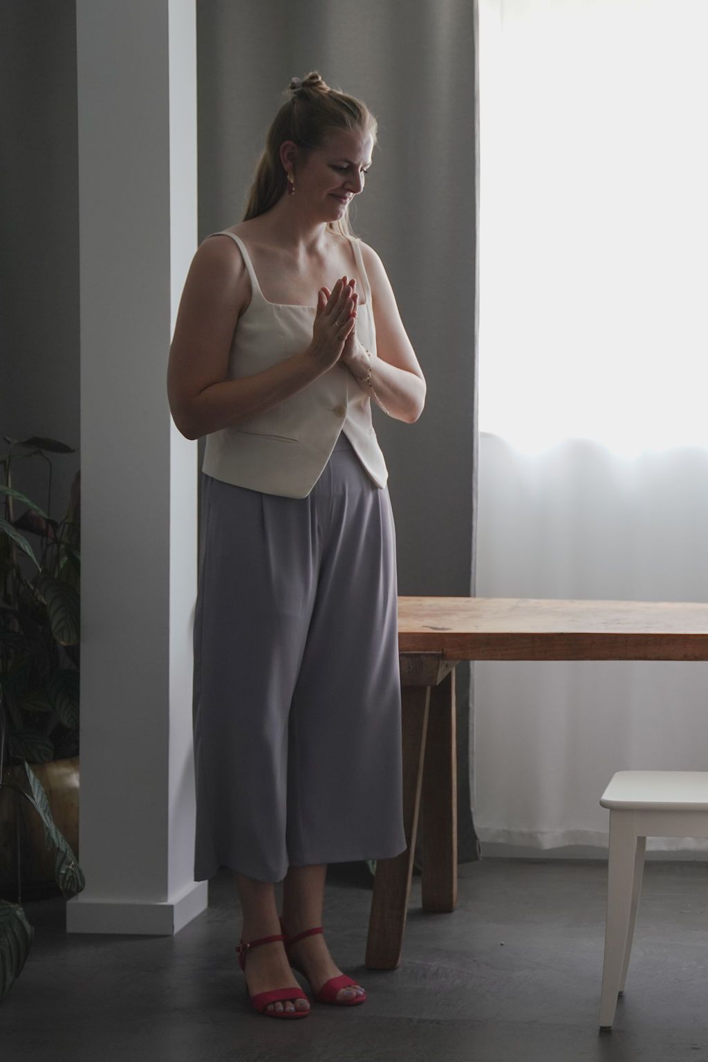 A person in a gray and white dress stands in a minimalist room with white walls and a wooden table.