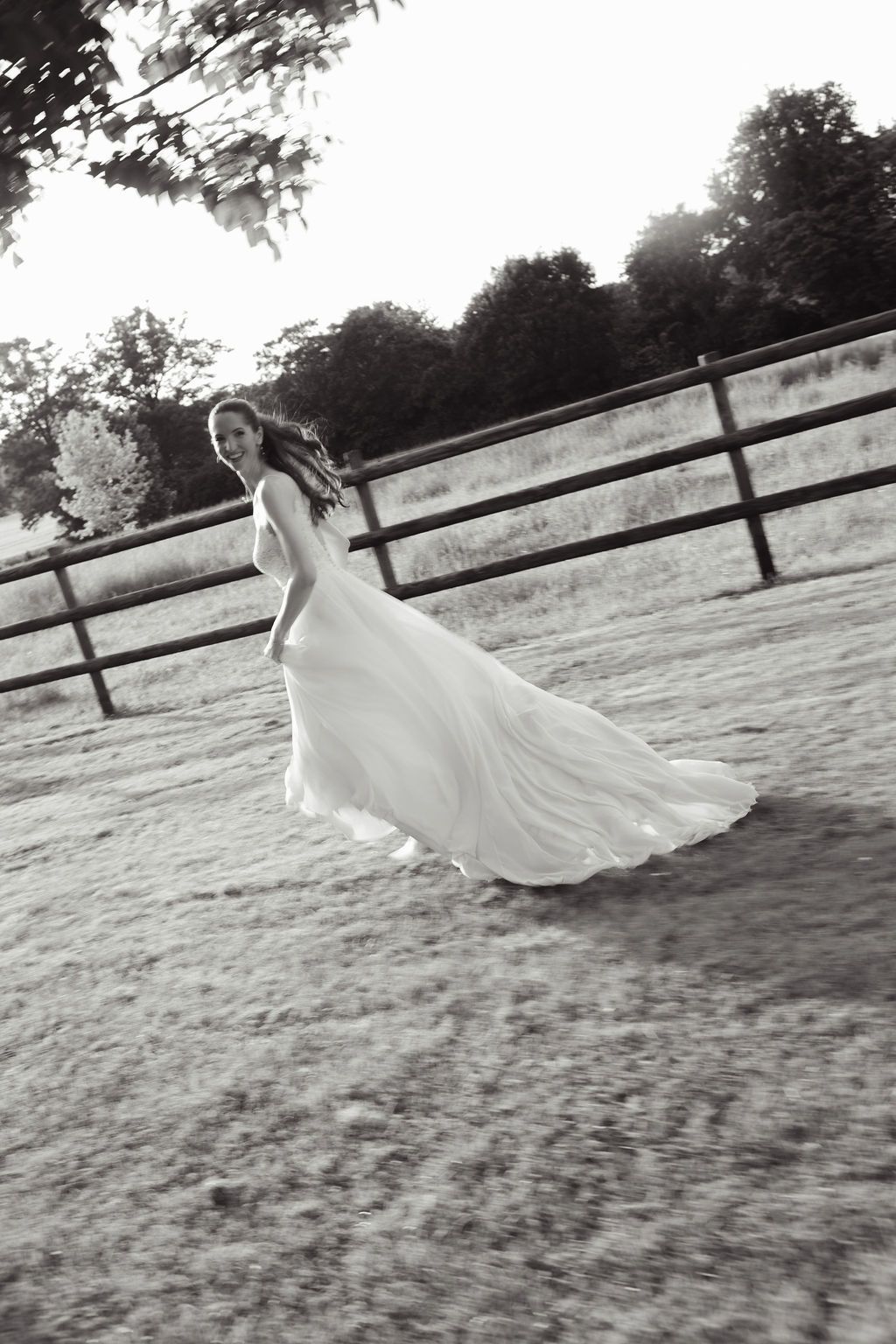 Bride in flowing white gown twirling in a rustic outdoor setting with wooden fence and trees in black and white photo.