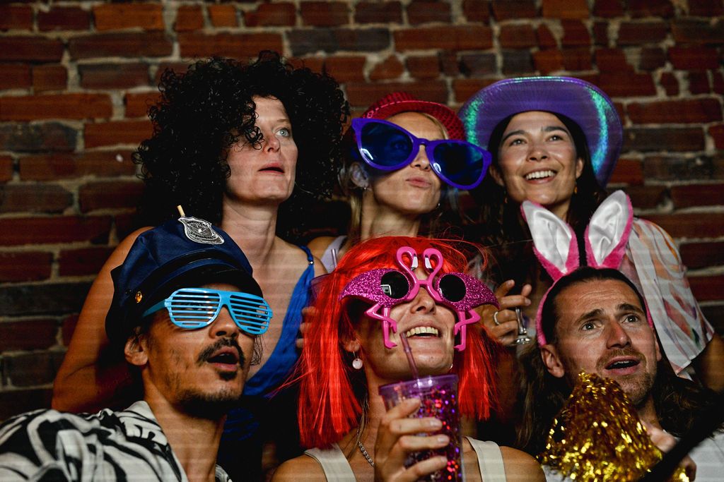 A crowd of people wearing colorful party hats, sunglasses and festive accessories smile and celebrate together.