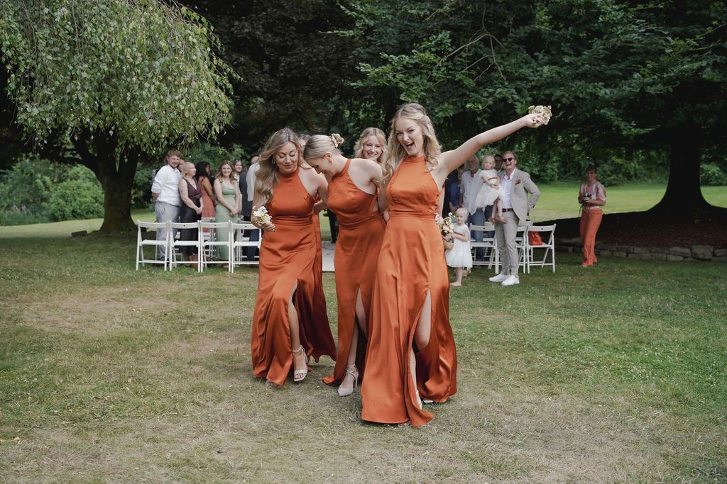 Wedding party members in orange dresses walk together on grass during an outdoor celebration.