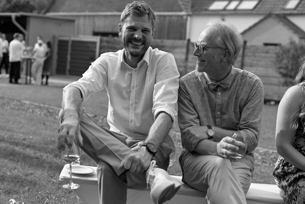 Two people sit together outdoors on a sunny day, smiling and engaged in conversation.