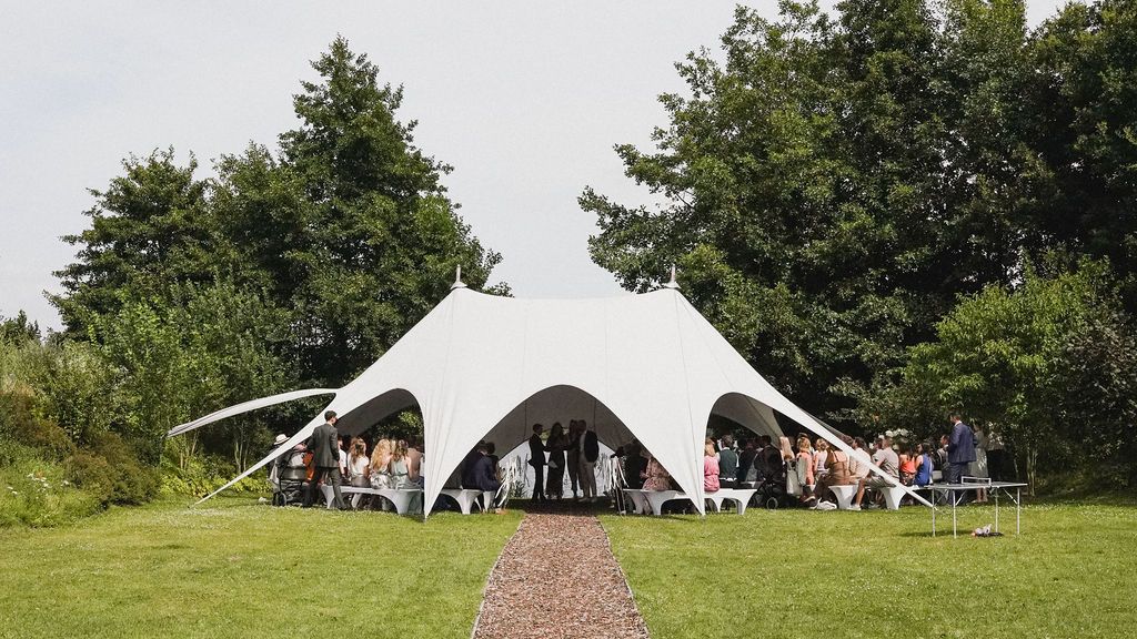 A large white event tent stands among green trees in a sunny outdoor setting.