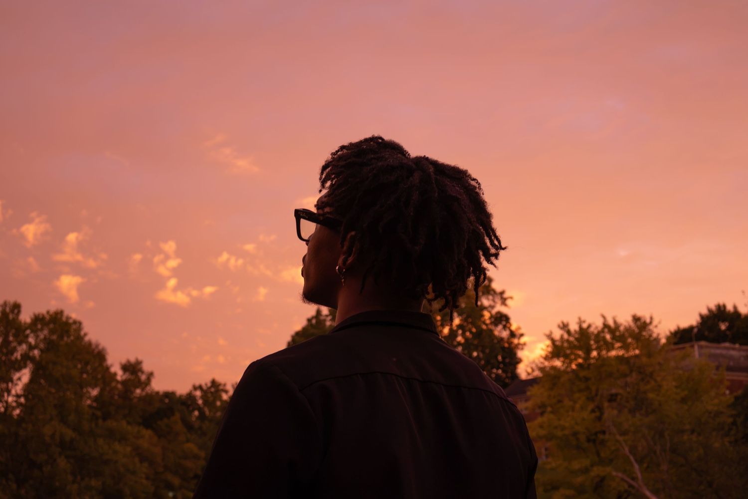 Silhouette of a man with dreadlocks and glasses against a vibrant pink and orange sunset sky.