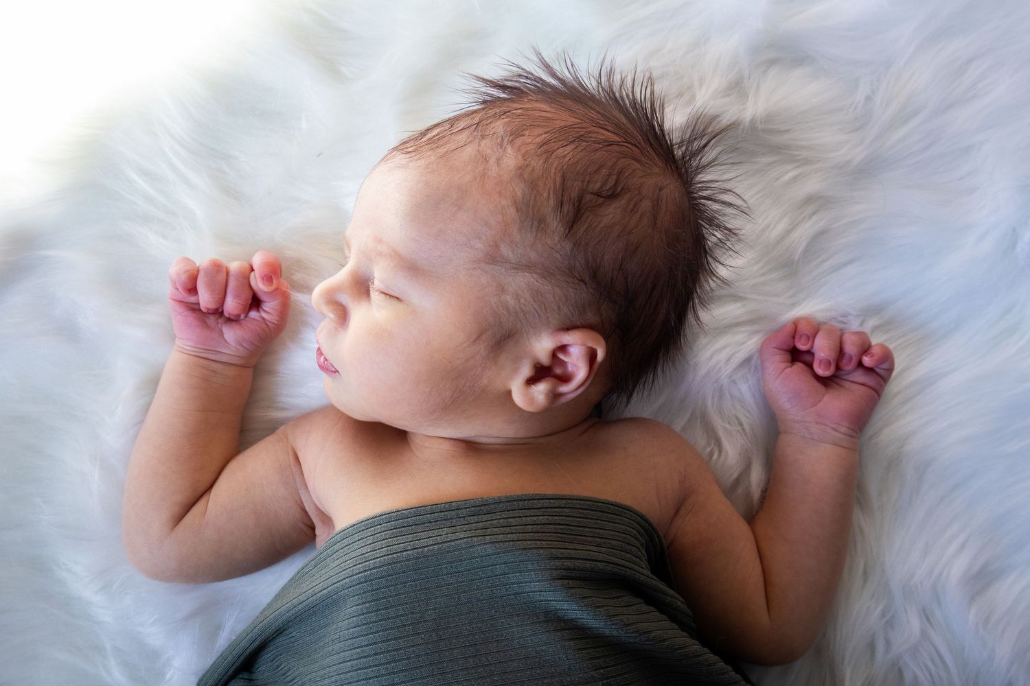 Peaceful newborn baby sleeping on white fluffy blanket, wrapped in green swaddle with tiny fists raised.