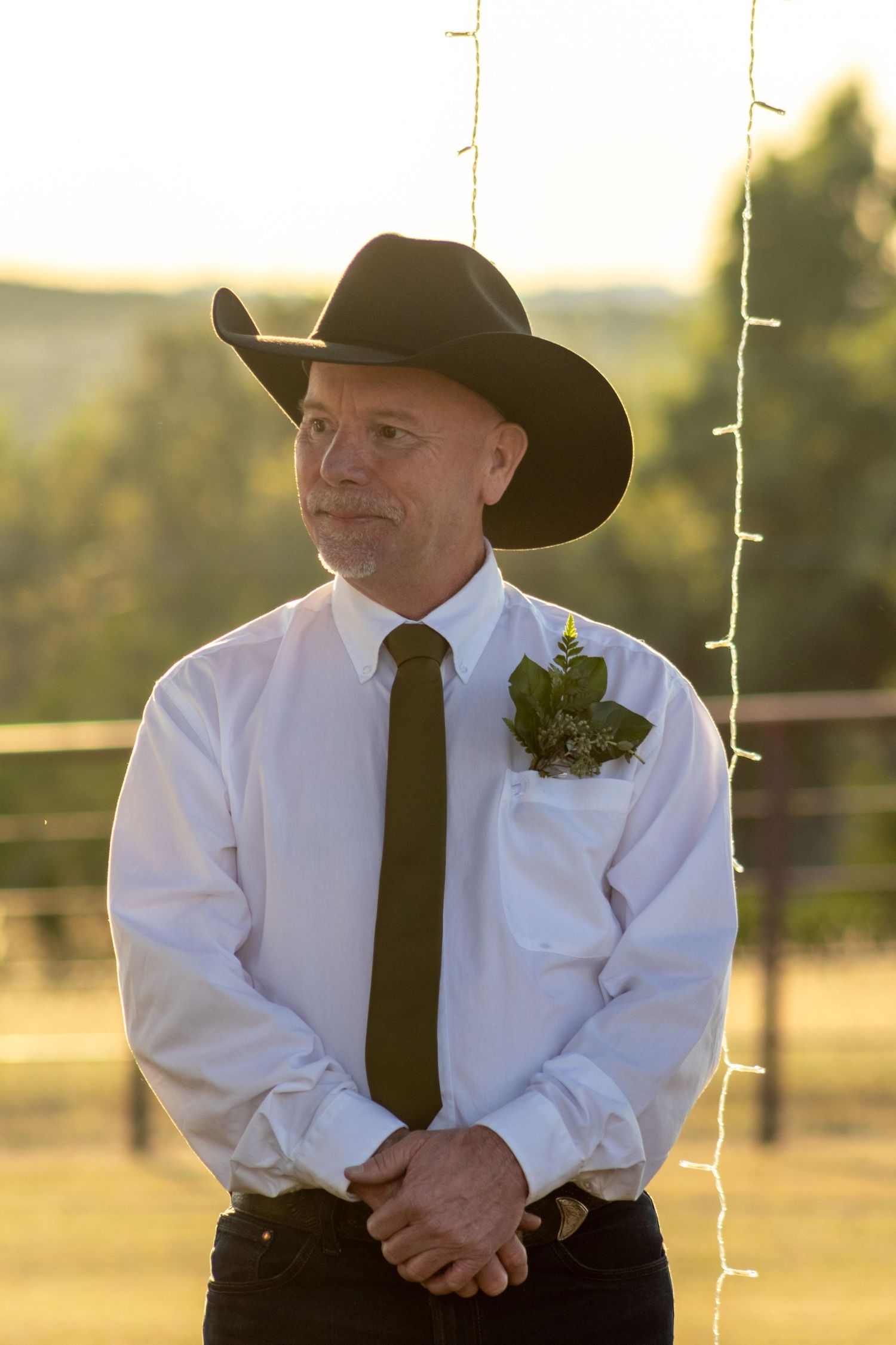 Man in black cowboy hat, white shirt, olive tie, and boutonniere at outdoor wedding.